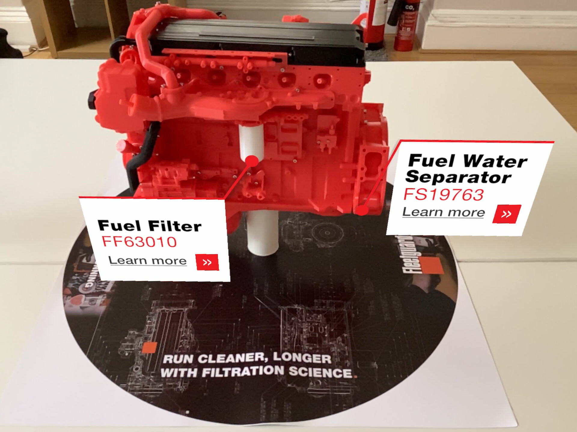 A red engine model on a circular Fleetguard display base with augmented reality labels pointing to the fuel filter and fuel water separator.