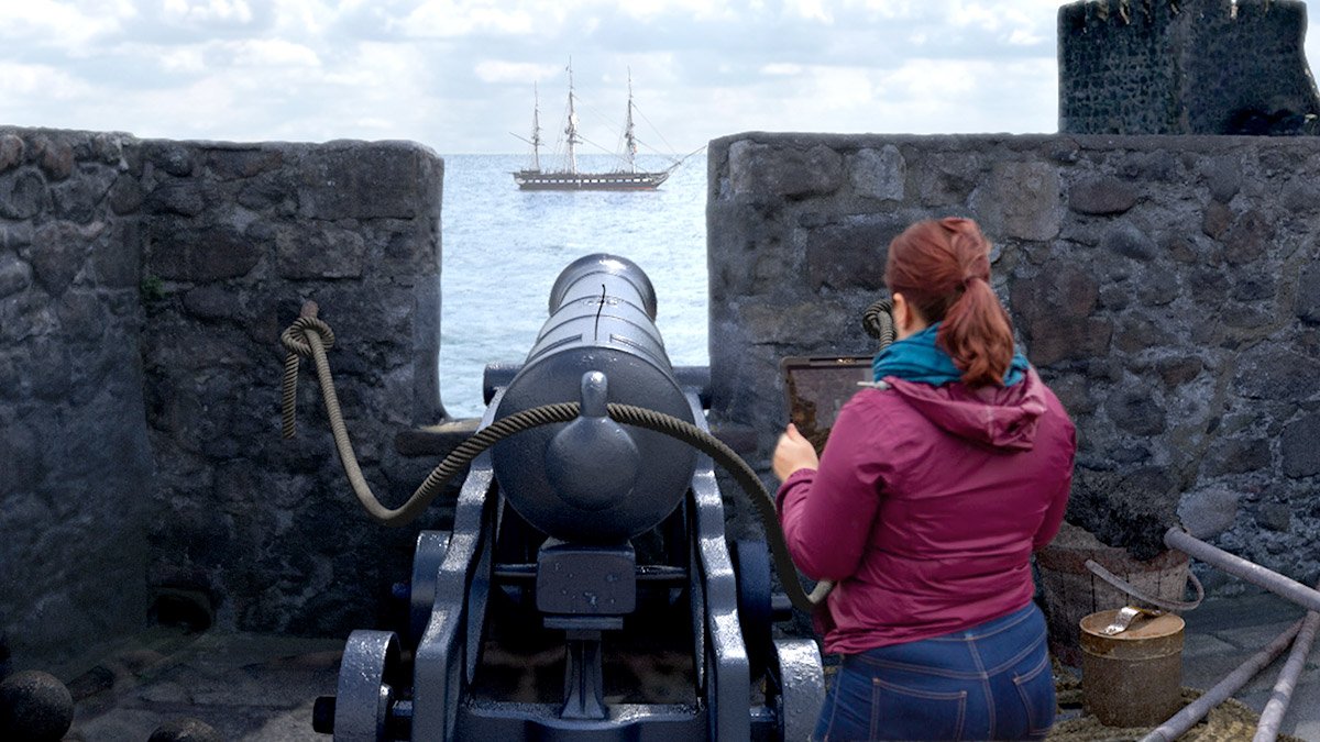 Person holding iPad in front of historical cannon testing CGI immersive AR heritage site reconstruction app for Carrickfergus Castle, Tourism Northern Ireland. The augmented reality scene as background