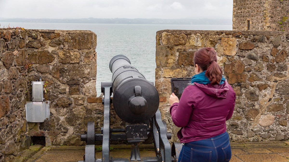 Person holding iPad in front of historical cannon testing CGI immersive AR heritage site reconstruction app for Carrickfergus Castle, Tourism Northern Ireland